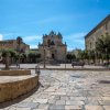 Piazza con chiesa storica, panchine e pavimentazione in pietra sotto un cielo blu.