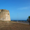 Torre in pietra vicino al mare su un prato arido, con un cielo blu sullo sfondo.