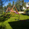 Parco giochi con altalene, scivolo, bilico e casetta, circondato da alberi.