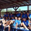 Un gruppo di persone in magliette blu sedute all'aperto su panchine di plastica sotto una tettoia.
