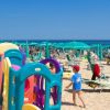 Spiaggia con ombrelloni verdi e area giochi colorata per bambini.