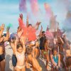 Gruppo di persone su una spiaggia lancia polveri colorate in aria, creando un'atmosfera festosa.