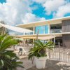 Edificio moderno con terrazza, sedie bianche e piante tropicali, cielo blu con nuvole.