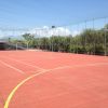 Campo sportivo all'aperto con pavimentazione rossa, circondato da alberi.